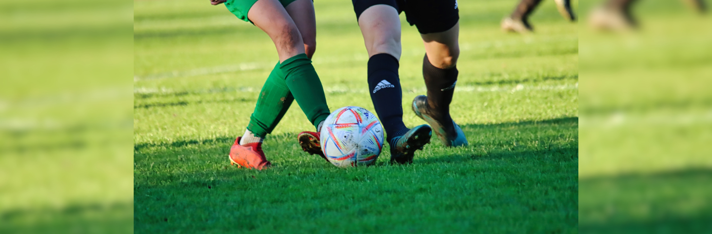 A football player in green shorts and socks and orange boots being dispossessed by a player in black shorts, socks and boots.