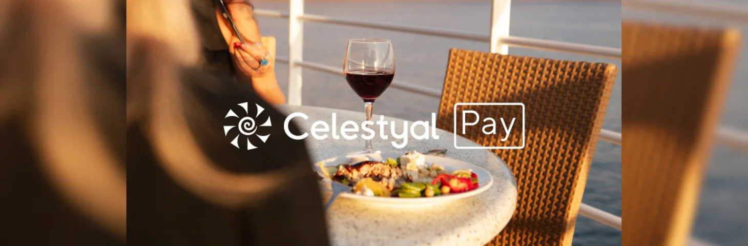 Two people sat down eating a meal with a glass of red wine on the deck of a cruise ship overlaid with the Celestyal Pay logo.